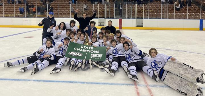 Group of hockey players and coaches on the ice at SNHU arena with state championship banner and trophy