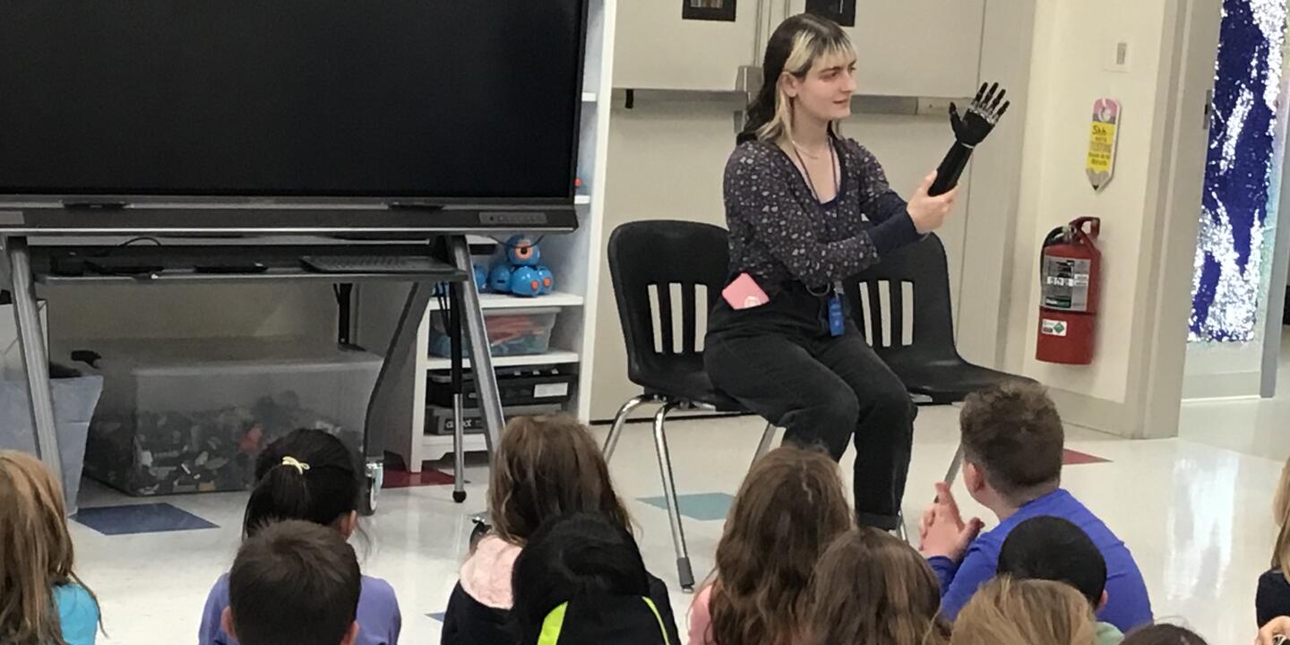 A woman sits in front of a room of second grade students explaining how her prosthetic arm and hand work.