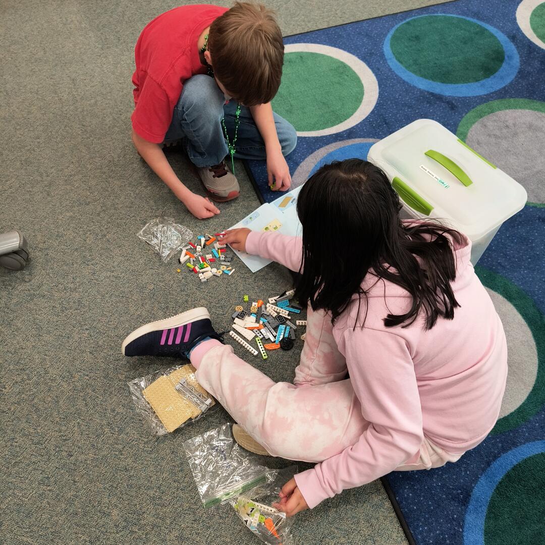 A boy and a girl sitting on a blue and green rug following a LEGO booklet on how to put together a LEGO creation.