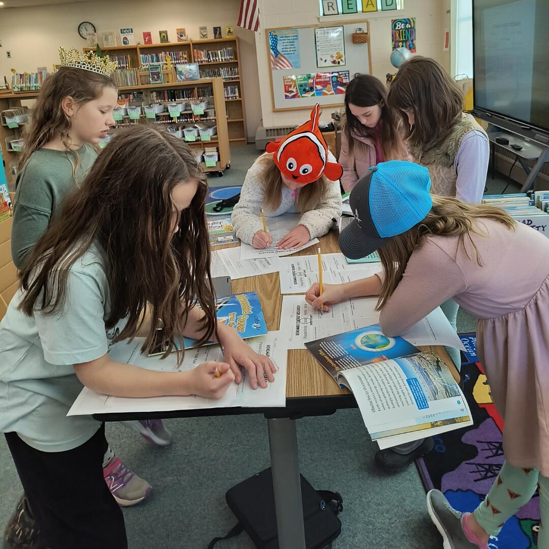 Six girls huddled around a table in a school library completing research notes.  One is wearing a silly, orange fish hat and another is wearing a bright, blue beanie.