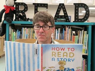 Man reading "How to Read a Story" in front of a shelf of books with The Cat in the Hat and the letters "READ" on shelf behind him.