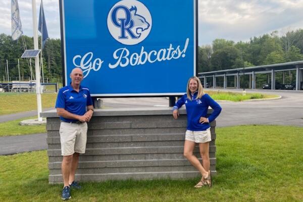 A photograph of Interim Principal Bill Sullivan and Interim Assistant Principal Alida Carter standing in front of Oyster River Middle School's "Go Bobcat" sign. Both are wearing blue Bobcat shirts.